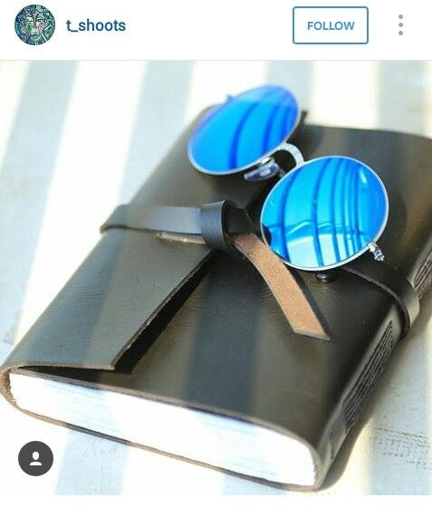 Jurnal kulit Hibrkraft berwarna gelap dengan tali pengikat, di atasnya diletakkan kacamata dengan lensa biru cerah.