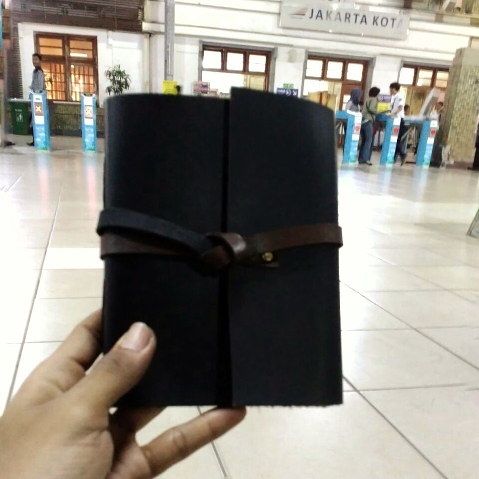 Jurnal kulit Hibrkraft berwarna hitam digenggam di dalam Stasiun Jakarta Kota yang bersejarah.