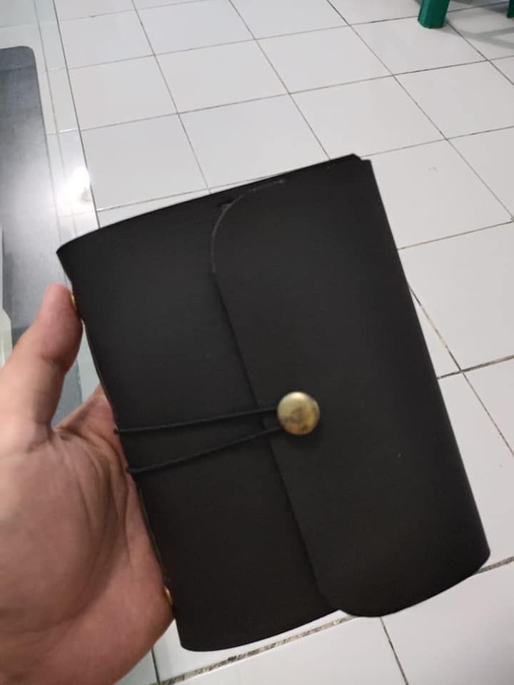 Jurnal kulit Hibrkraft A6 berwarna hitam dengan kancing kuningan, digenggam erat di tangan.