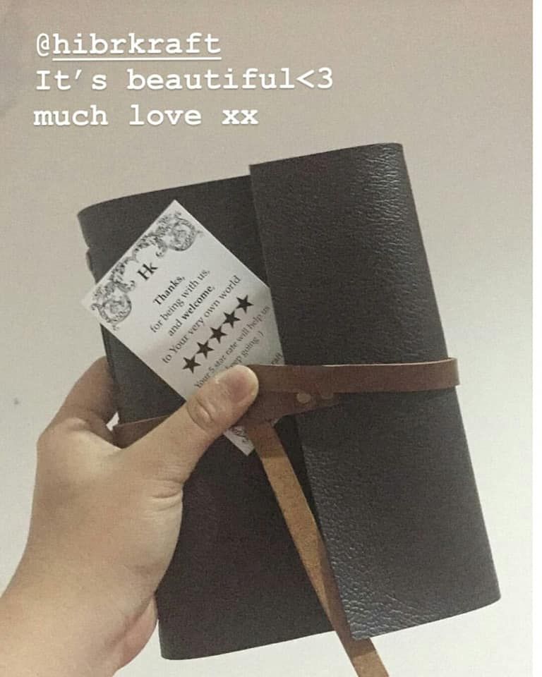 Sebuah tangan memegang jurnal kulit Hibrkraft berwarna gelap, dengan kartu ucapan terima kasih terselip di tali pengikatnya.