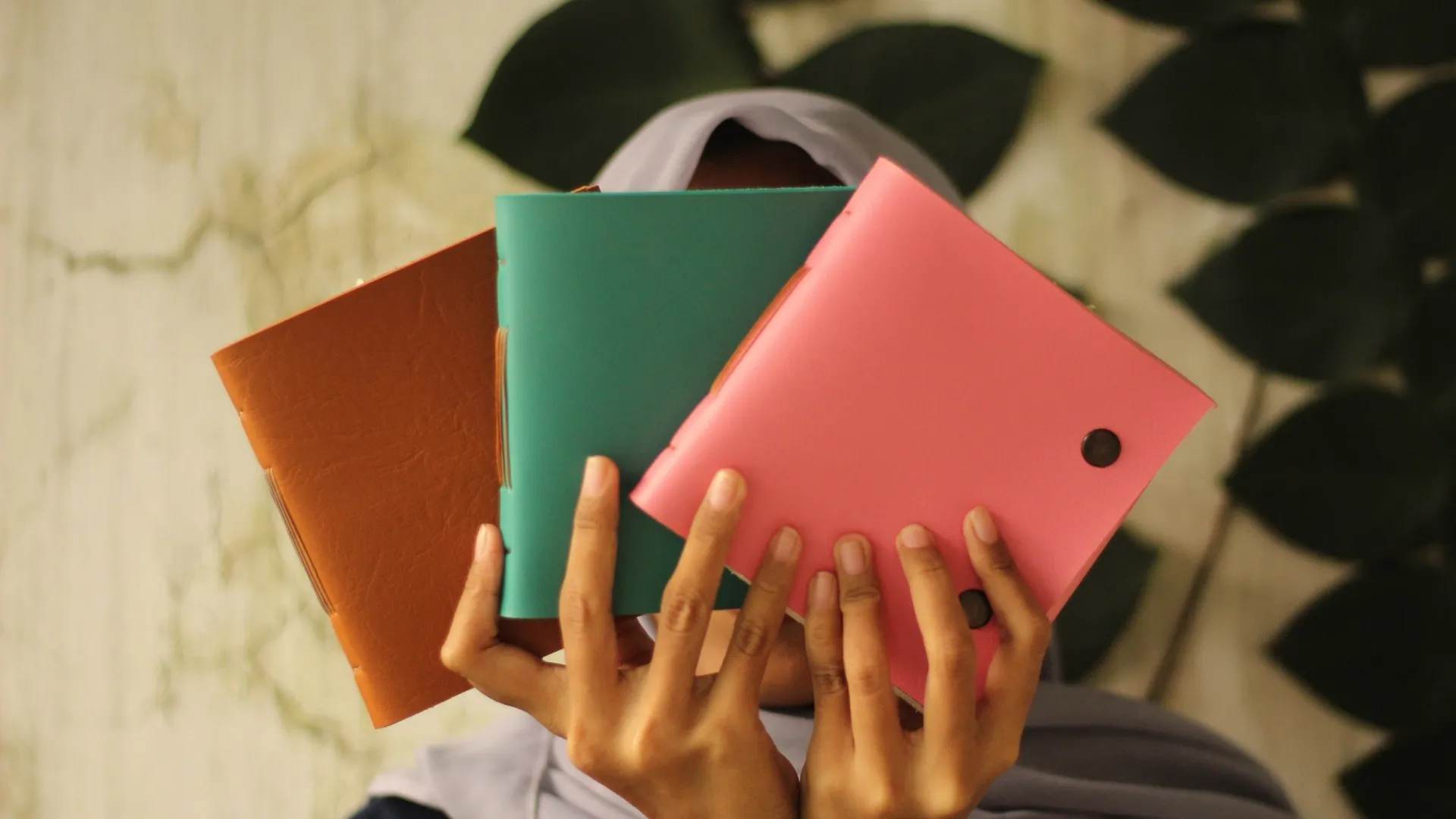 Seseorang memegang tiga jurnal kulit dengan warna berbeda: coklat, hijau toska, dan pink, menunjukkan variasi dan personalisasi.
